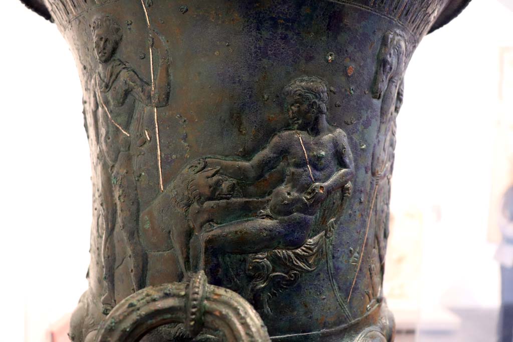 IX.13.1-3 Pompeii. February 2021.
Bronze krater with scenes in relief, found in triclinium, on display in Antiquarium, VIII.1.4.
Detail of the second sitting figure, young and naked, caressing an animal with one hand and holding a sword by the hilt in the other.