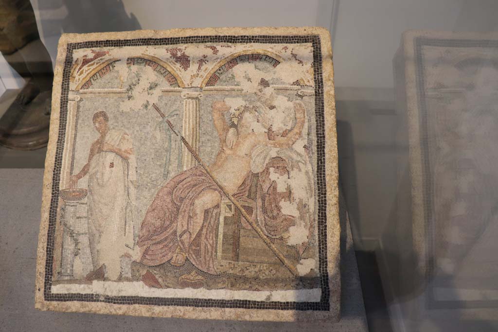 Via del Vesuvio Pompeii. February 2021. Mosaic on display in Pompeii Antiquarium.
Central mosaic picture that adorned a fountain in a house on Via del Vesuvio, in front of which there was a statuette of a rabbit.
Photo courtesy of Fabien Bièvre-Perrin (CC BY-NC-SA).
According to Kuivalainen, this was the centre mosaic below a niche with columns and a pediment from V.6.7. 
The mosaic consists of 3 figures in an architectural setting against a light blue background and white floor area.
On the left stands a man holding his right hand over and looking down on a brownish basin on top of a low separate stand, a white column, entwined with a garland.
The main figures are a couple loosely embracing each other, a reclining drunken youth looking upwards towards a female figure, seated behind, higher up on the podium, looking down at him.
They are without doubt Bacchus and Ariadne.
See Kuivalainen, I., 2021. The Portrayal of Pompeian Bacchus. Commentationes Humanarum Litterarum 140. Helsinki: Finnish Society of Sciences and Letters, D16: p. 138-9.
