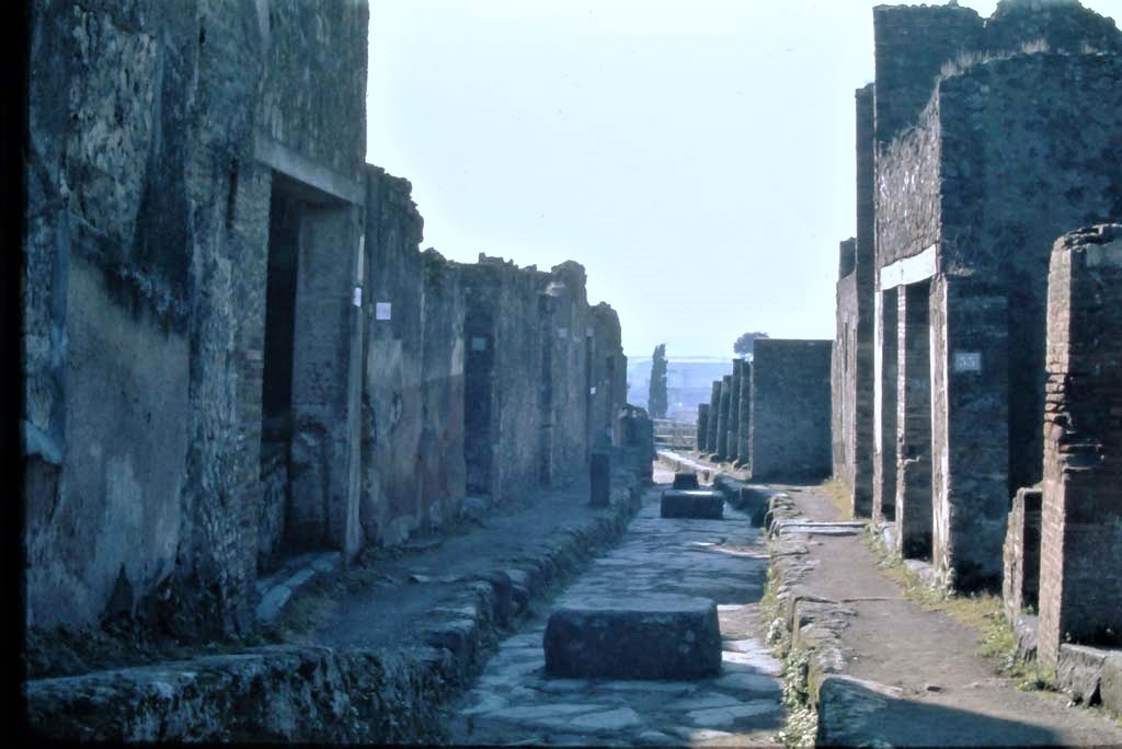 Vicolo dei Soprastanti, Pompeii. 4th December 1971. Looking west between VII.7 and VII.6.
On the left is the doorway to the bar at VII.7.18 followed by Vicolo el Gallo, and then doorway at VII.15.12.
On the right is the doorway of VII.6.33.
Photo courtesy of Rick Bauer, from Dr. George Fay’s slides collection.
