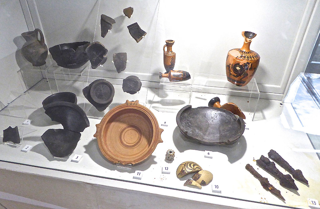 Santuario extraurbano del Fondo Iozzino. June 2017. More finds from site. Now in PAP deposits.
Photo courtesy of Michael Binns.
The exhibition information card identifies these as:
1: Campanian bucchero kantharos with Etruscan inscription, “mi marmarces tetanas” = “of Marmarce Tetana”, second half of 6th century BC
2: Campanian bucchero kantharos with Etruscan inscription, second half of 6th century BC
3: Campanian bucchero bowl with graffito bird, second quarter of 6th to 1st quarter of 5th century BC
4: Bucchero chalice with fan motifs, end of 7th to 1st quarter 6th century BC
5: Bucchero olpe, second half of 6th to 1st quarter of 5th century BC
6: Campanian bucchero bowl, second half of 6th century BC
7: Campanian bucchero kantharos, second half of 6th century BC
8: Attic black figure lekythoi with palmette decoration, circa 480 BC
9: Attic black figure lekythoi with hoplites, of 6th to beginning of 5th century BC
10: Corinthian aryballos decorated with hoplites, 580 to 560 BC
11: Bowl with painted decoration, archaic period
12: Faience bead, archaic period
13: Iron arrow heads, 6th century BC
14: Sherds of bucchero kantharoi and cups with graffiti, 6th century BC