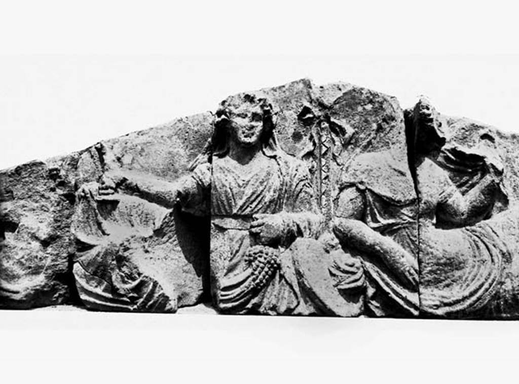 Tempio dionisiaco in località Sant’Abbondio di Pompei. The two gods on the tufa pediment from front of temple.
Photo courtesy of Ruth Bielfeldt.
The depiction shows a Dionysian scene of pyramidal symmetric structure. Shoulder on Shoulder lie the pair of gods in the centre, flanked each with a satellite scene, which extends from the edges of the gables
In the centre, between the gods, is the towering thyrsus-pole in the gable vertex and leaning upright against the thyrsus is a Tympanum. The characterization of the god as Dionysus exists because of the attributes, the grape in the left, the Kantharos in the right, the ivy wreath and the hair band. But the garment is unusual for the usually lightly dressed Dionysus. The god is in an ankle-length, highly girt, long-sleeved “Theaterchiton” like Apollo Citarista, but also Dionysus wears, for instance in representations of the Indian Triumph on imperial sarcophagi. Without comparison in Dionysian iconography is, however, the veiling of the back of the head through the coat, a costume feature that in connection with the strict frontality and the motionless posture, gives the God a hieratic aura.
The companion lying to the left of God, a youthful goddess of restrained erotic beauty, is more moved towards the opposite. In a spacious unveiling gesture, she pulls with the left, formerly raised, hand the veil laid over the back of the head, to the side. Only the left part of her head remains, with the wavy hair, parted in the middle. The face is mostly broken away, as the block joint ran right through the face. A sign of their physical attractiveness is the identical motif of the chitons slipped off the shoulder, which frees the unclothed shoulder, and the rich jewelry, a torque around the neck and an upper arm band on the left arm. However, they are not specific attributes to identify her.
See Bielfeldt R., Der Liber-Tempel in Pompeji in Sant’Abbondio. Oskisches Vorstdtheiligtum und kaiserzeitliches Kultlokal, dans MDAI-Römische Abteilung, 113, 2007, pp. 323-4, Abb. 5, 6.