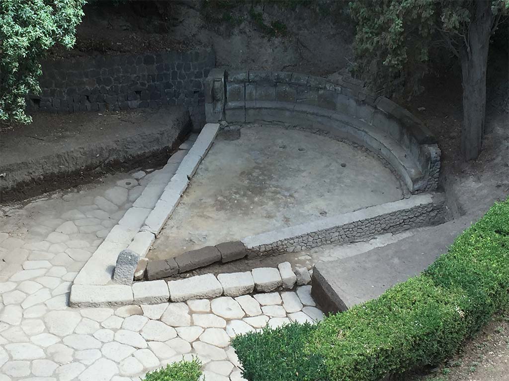 NGH Pompeii. 2015. Schola tomb after clearance showing previously covered tomb and basalt block roads.
The continuation of ancient Via di Nola into unexcavated area is on the left.
The area immediately outside of Porta Nola (the Nolan Gate) was first cleared in 1907–8.
Since then, soil had once again accumulated opposite the gate, re-burying the tomb.
In 2015 the Porta Nola Project has cleared the basalt block roads and brought the monument back to light.
Excavation inside the monument in front of the benches revealed a thick layer of floor preparation for the pavement which has since disappeared.
See BSR Pompeii Project Porta Nola Necropolis 2015 Excavations