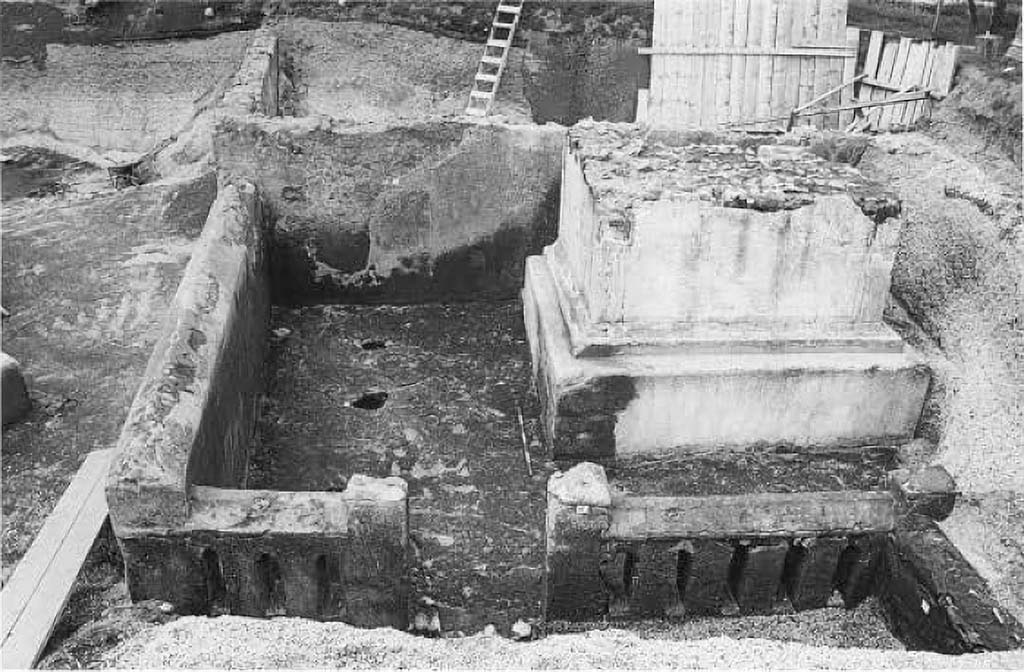 Pompeii Porta Sarno Necropolis. 1999. Podium tomb A. Funerary site with a monument on podium and enclosure.
In 1999 a new necropolis area was also discovered in which, in particular, is a large funerary monument - of the "a dado" (cube) type on a podium - of which, unfortunately, the upper part has been lost with it the epigraph that probably must have been there, so that the owner remains unknown. The building, in opus incerta, with corners in brick work, covered with white plaster, is contained in an enclosure in opus incerta work, in which narrow vertical windows open. At the base of the rear side, there is a niche, in turn containing a smaller niche in which a columella with the related cinerary material was placed, stuck in the ground. In the space between the enclosure and the podium, cavities relevant to the roots of trees, probably cypresses, that surrounded the monument were highlighted.
Nel 1999 è stata, inoltre, scoperta una nuova area di necropoli nella quale è da segnalare, in particolare, un grande monumento funerario - del tipo "a dado" su podio - del quale, purtroppo è andata perduta la parte superiore e con essa l'epigrafe che, verosimilmente, doveva esserci, così che ne resta sconosciuto il proprietario. L'edificio, in opera incerta, con ammorsature angolari in opera laterizia, rivestito di intonaco bianco, è inserito in un recinto in opera incerta, nel quale si aprono strette afinestrature verticali. Alla base del lato posteriore, si apre una nicchia, a sua volta contenente una nicchia più piccola nella quale era posta, infissa nel terreno, una columella con il cinerario relativo. Nello spazio tra il recinto e il podio, sono state evidenziate cavità pertinenti alle radici di alberi, probabilmente cipressi, che circondavano il monumento.
See/Vedi Suburbio Orientale in Rivista di Studi Pompeiani X, 1999, p. 181, fig. 2.
