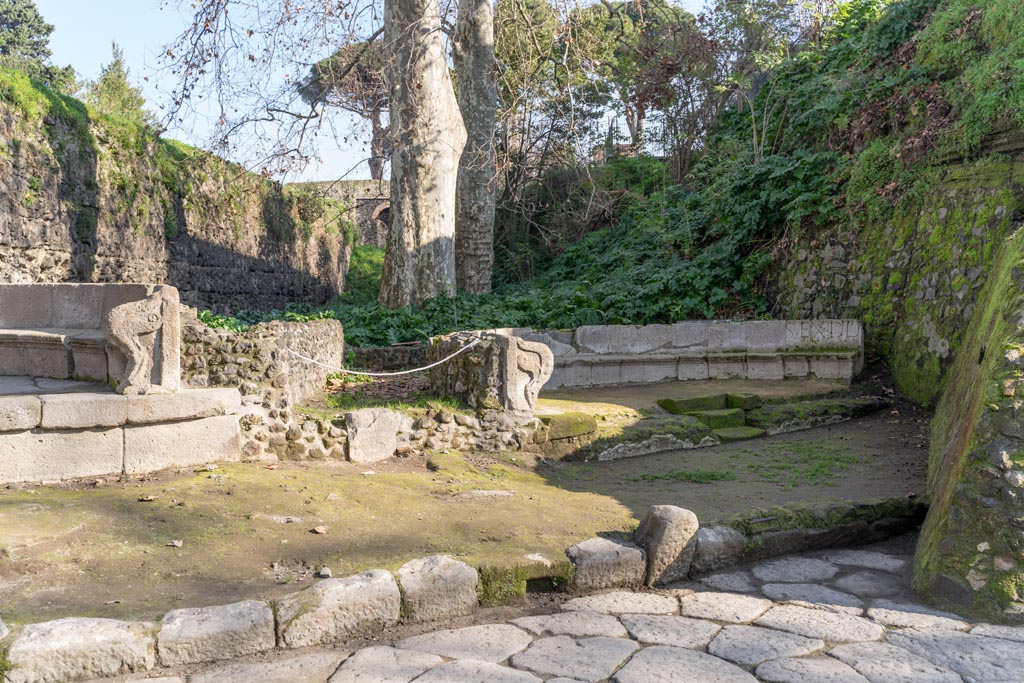 SGD Pompeii, south end on left. January 2023. Looking east towards two schola tombs, with SGF, on right.
The entrance to the enclosure behind the tomb is in the centre.
In front of the entrance the road surface, a drain set in the kerb, and the steps in the centre front of the tomb, are visible.
Photo courtesy of Johannes Eber.