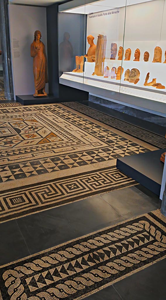 Villa Urbana, Varano, Stabiae. July 2019.
Mosaic floor as seen in room on First Floor in Naples Museum.
Photo courtesy of Giuseppe Ciaramella.
According to Pisapia –
“Of the four rooms that were paved with mosaic floors, the designs that La Vega made of two of these, lost, nos, 122 and 123, are shown in fig.19, while Ruggiero states that the other two are in Real Museo Borbonico and pave the first and second rooms of the Santangelo Collection.
Of this information, however, only part is correct, since, in the first room of the aforementioned Collection, it has been recognised that there is a mosaic from Lucera, while in the second room it is the floor that we indicate as no.124.”
(Dei quattro ambienti che erano pavimenti a mosaico si riporta alla fig.19 i disegni che il Vega fece di due di essi n.122 e 123 perduti, mentre il Ruggiero afferma che gli altri due sono al Real Museo Borbonico e pavimentano la I e la II sala della Collezione Santangelo.
Di questa notizia, però, soltanto parte era esatta, poiché, nella I sala della Collezione citata e stato riconosciuto esservi un mosaico da Lucera, mentre nella II sala e il pavimento che indichiamo con il no.124).
See Pisapia, M. S. 1989, in Mosaici antichi in Italia, Regione prima. Stabia, Roma, (p.67-70, no.124).
According to Ruggiero –
“Di questo edificio Avanza solamente una copia della pianta (senza il riscontro dei numeri relativi al Giornale perduto) e i musaici dei pavimenti di quattro stanze disegnati dal la Vega; due dei quali si pubblicano con la detta pianta nella Tav. XVIII e due altri si tralasciano, potendosi vederli nel Museo nazionale, l’uno nel mezzo della prima sala dei vasi e l’altro parimente nel mezzo della seconda sala della collezione Santangelo.”
(Of this building remains only a copy of the plan (without the confirmation of the numbers relating to the lost Giornale) and the mosaics from the floors of four rooms drawn by La Vega; two of which are published with the said plan in Tav. XVIII and two others have been left out, as they can be seen in the National Museum, one in the middle of the first room of vases and the other equally in the middle of the second room of the Santangelo collection.)
See Ruggiero, M. (1881). Degli scavi di Stabia dagli 1749 al 1782. (p.356).