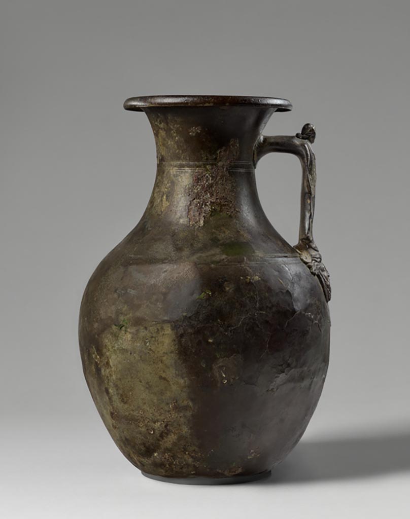 Discovered in a villa in Boscoreale. Bronze amphora with standing female figure and satyr mask.
Digital image courtesy of the Getty's Open Content Program. Now in the Getty Museum, inventory number 72.AC.143.
Aside from two pairs of incised lines on the shoulder and neck of the vessel, the handle, which takes the form of a standing female figure, provides the only decoration on this amphora (storage jar). She wears an ankle-length garment and mantle and holds her arms at her sides. The attachment plate upon which she stands is shaped as a mask depicting a bearded satyr wearing a fillet.
Satyrs were frequent companions of the wine god Bacchus, and vessels of this type were often used to hold wine at banquets. This amphora was discovered in a villa at Boscoreale or in the vicinity, although the exact findspot is unknown. The owners of the luxurious homes in this area used finely shaped vessels decorated with a variety of images, including mythological figures, theatre masks, animal heads and feet, and floral ornaments.