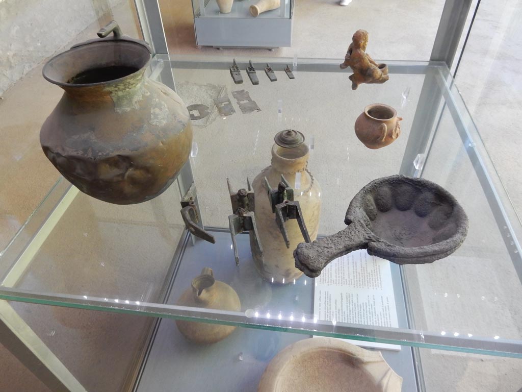 Complesso dei triclini in località Moregine a Pompei. May 2018. Finds on display in Pompeii Palaestra.
Bronze jug with handle in the form of a thumb.
Rectangular and circular bronze plaquettes of locks with holes for fixing on the wooden support.
Bronze elements of locks where the keys were inserted.
Circular bronze nail for furniture.
Two iron baking moulds.
Thin-walled ceramic jar of Vesuvian production decorated with a human face.
The ithyphallic glazed clay lamp in the form of a satyr at the rear right is from Edifici B.
Photo courtesy of Buzz Ferebee.
See Nappo, S. C., 2012.: Un esempio di architettura ricettiva alla foce del Sarno in Rivista di Studi Pompeiani XXIII, p. 97 fig. 27.