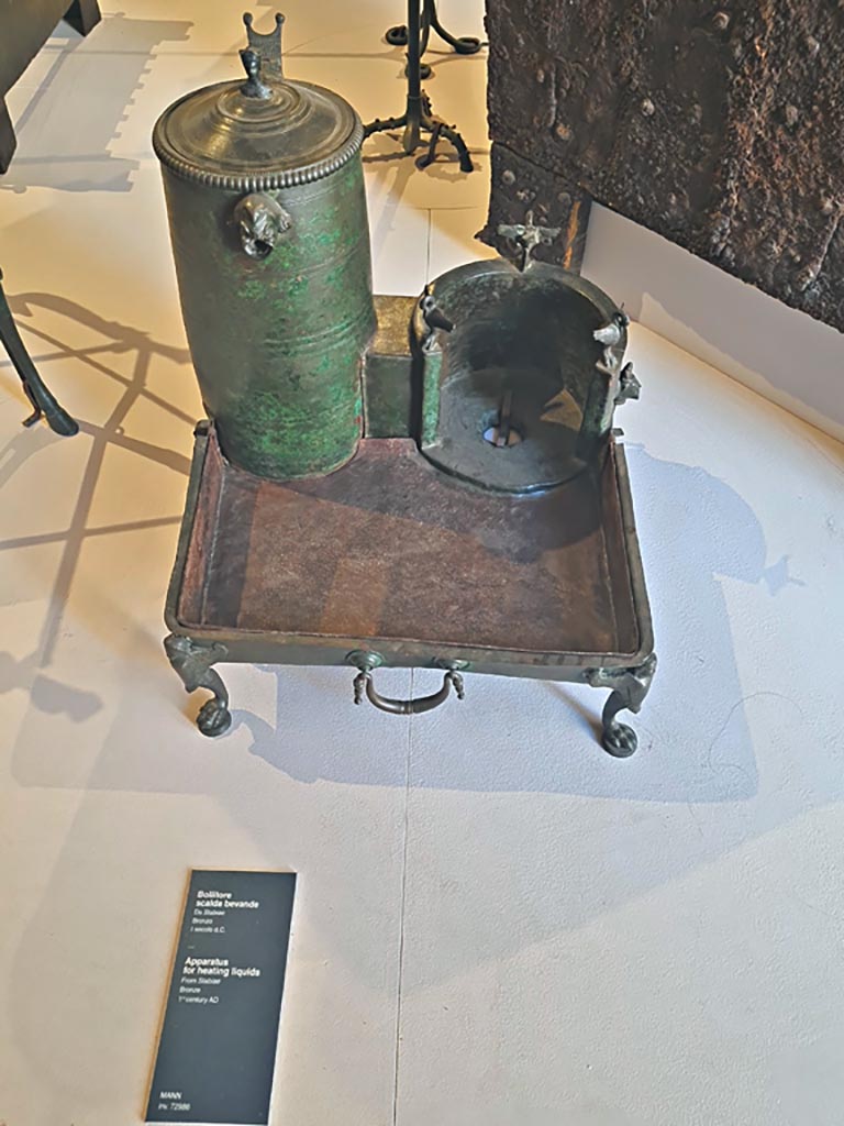 Stabiae, Villa Arianna, October 2023. Room 10, heating appliance, inv. 72986. Photo courtesy of Giuseppe Ciaramella.
On display in “L’altra MANN” exhibition, October 2023, at Naples Archaeological Museum.
According to Pagano and Prisciandaro, this was found in room 10, 23rd October 1758.
Their other references were Rami inediti, fig.198 a and b, Ruesch,1695, Tav. IV, Masseria Irace, 10.
See Pagano, M. and Prisciandaro, R., 2006. Studio sulle provenienze degli oggetti rinvenuti negli scavi borbonici del regno di Napoli. Naples: Nicola Longobardi,
(p.242).
Sulla destra del fornello un rubinetto, decorato a testa di leone, permetteva al liquido riscaldato di uscire secondo le esigenze. L’ansa del coperchio è a forma di bustino di Mercurio e sulla parete del contenitore è fissata una applique a maschera comica; tre cigni decorano il fornello e tre maniglie “a goccia” sono applicate ai lati del vassoio.
(trans: On the right of the stove a tap, decorated with a lion’s head, allowed the heated liquid to flow out as needed. The handle of the lid is in the shape of a head of Mercury and fixed on the wall of the container is a comic mask. Three swans decorate the stove and three “teardrop” handles are applied to the sides of the tray.)
See Le Collezioni del Museo Nazionale di Napoli, 1986, Rome, De Luca Editore, (p.182-3, no.74), and with reference to MB 1829, V, Tav. XLIV.