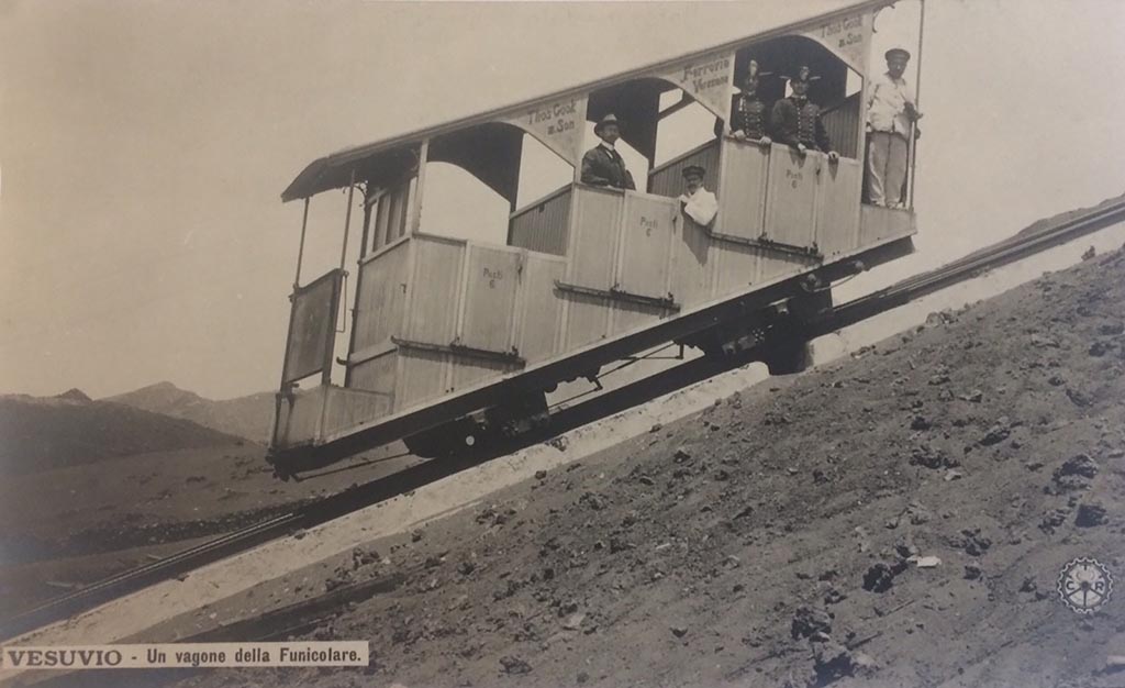 Postcard showing a Wagon of the Vesuvius Funicular, with handwritten note on rear- “The 1904 Funicular only lasted 2 years”.
Photo courtesy of Rick Bauer.
This photo must be dated between 1904 when the cars were introduced and 1906 when they were destroyed..
The funicular was modernised at the same time as Cook’s Pugliano to Vesuvius light railway was opened in 1904.
The monorails had been removed and the track was now a one metre gauge single track with a central passing loop.
These cars were new in 1904 and seated 18 with 6 standing.
Thomas Cook and Son’s name is on the upper part of the structure. The wording on each of the three doors is “Posti 6” (6 places).
Two years later, in a tremendous eruption, on 7 and 8 April 1906 the lower and upper station, the equipment, the machinery, the two funicular cars were destroyed.
Everything was buried under a 20-30m high ash blanket.
Although the light railway as far as the hotel Eremo was quickly dug out, the funicular had to wait.
Only in 1909, according to the project of the engineer Enrico Treiber, were the works for a new funicular ended and it reopened in 1910.