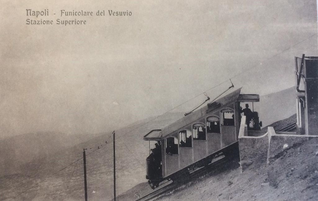 Postcard for Vesuvius Funicular, showing Upper Station. Not dated but must be between 1910 and 1944.
There is a handwritten note on rear – “The 1910 Funicular lasted until 1944”. Photo courtesy of Rick Bauer.
After the 1906 eruption the funicular was out of action and reconstruction was only completed in 1909.
It reopened in 1910 with two new 5-bay end loading cars, numbered 1 and 2, which seated 16 with 8 standing.
The car in this photo is car number 1.
The line now had overhead wire and the cars bow collectors to provide electric light, bell signals and a telephone.
The funicular could now run an after dark service.
The funicular was permanently closed after being destroyed by the Vesuvius eruption of 1944.