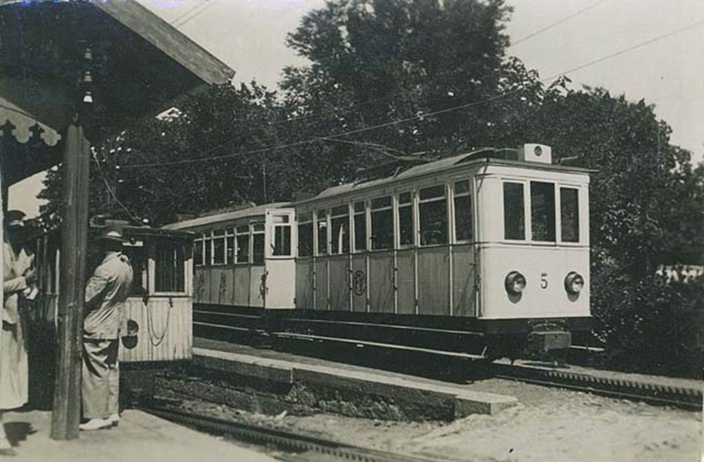 Vesuvius to Pugliano railway. 1934. Hotel Eremo station. Photo courtesy of Rick Bauer.
The same carriages were used for a journey along the whole of the track.
From Pugliano to the power station and from Eremo to the Funicular the trains used their own power and traction was by simple adhesion to the rails.
The rack system of the track is visible in front of the rack locomotives and carriages.
The rack locomotive on the left was used to push the carriages up the steepest part of the track with up to 25% gradient from the power station to Eremo.
Traffic had developed to the point where larger cars were needed.
Car number 5 on the right was introduced in the 1920’s and seated 34 passengers who were fully enclosed.
Two other newer cars, numbered 6 and 7 each seated 44 passengers.
The original cars 1 to 3 had seated only 24 people with an additional 6 standing on the platform. They were open above the waistline with curtains for use against wind and rain. A fourth enclosed car had been added when the railway was extended to the relocated and rebuilt funicular base station on 6th January 1913.
See Smith P, 1998. Thomas Cook and Son’s Vesuvius Railway in Japan Railway and Transport Review March 1998.